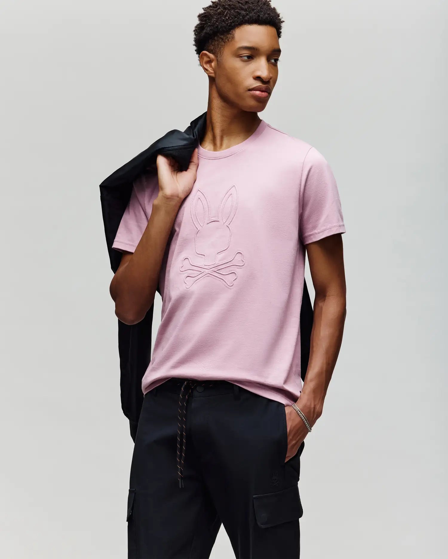A young man poses against a light gray background, holding a black jacket over his shoulder and wearing Psycho Bunny's MENS REMY GRAPHIC TEE (MT0200059) in light pink, featuring an embossed Bunny logo and crossbones design, paired with black pants. (4)