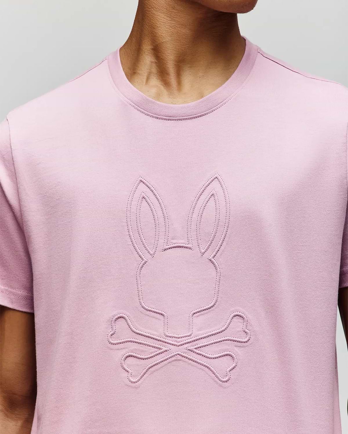 A person wears a Psycho Bunny MENS REMY GRAPHIC TEE (MT0200059) in light pink, crafted from soft Pima cotton, with an embossed bunny logo above two crossed bones on the front. The person’s face is not visible. (3)