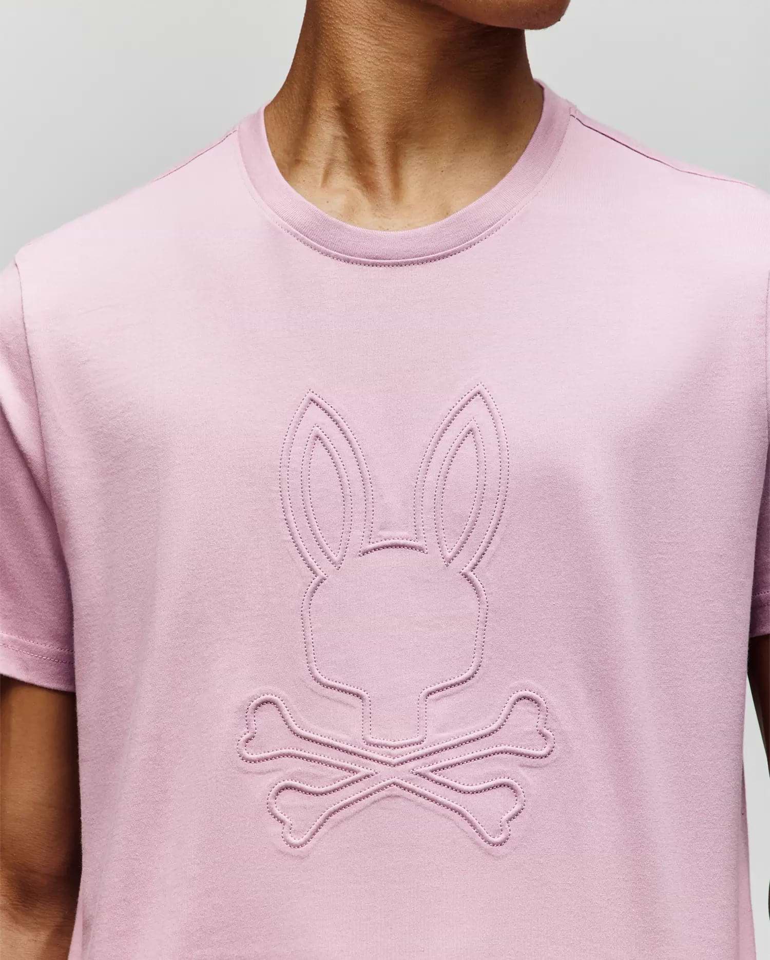 A person wears a Psycho Bunny MENS REMY GRAPHIC TEE (MT0200059) in light pink, crafted from soft Pima cotton, with an embossed bunny logo above two crossed bones on the front. The person’s face is not visible. (3)