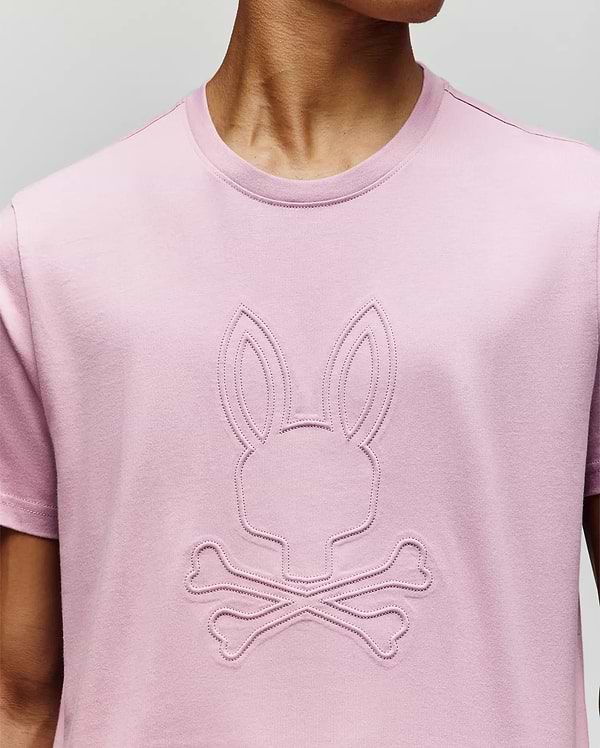 A person wears a Psycho Bunny MENS REMY GRAPHIC TEE (MT0200059) in light pink, crafted from soft Pima cotton, with an embossed bunny logo above two crossed bones on the front. The person’s face is not visible. (3)