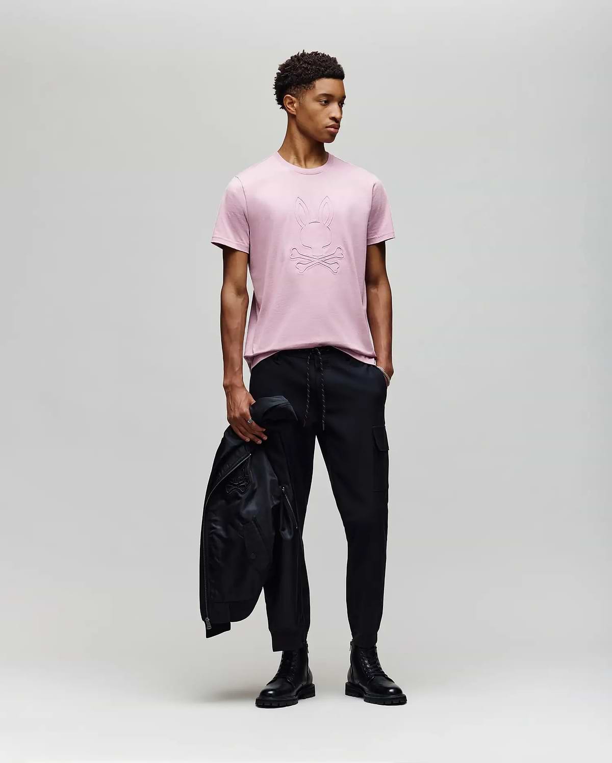 A young man stands against a plain background wearing a light pink Psycho Bunny MENS REMY GRAPHIC TEE - MT0200059 made from soft Pima cotton, paired with black pants and boots, holding a black jacket in his left hand while looking right. (5)