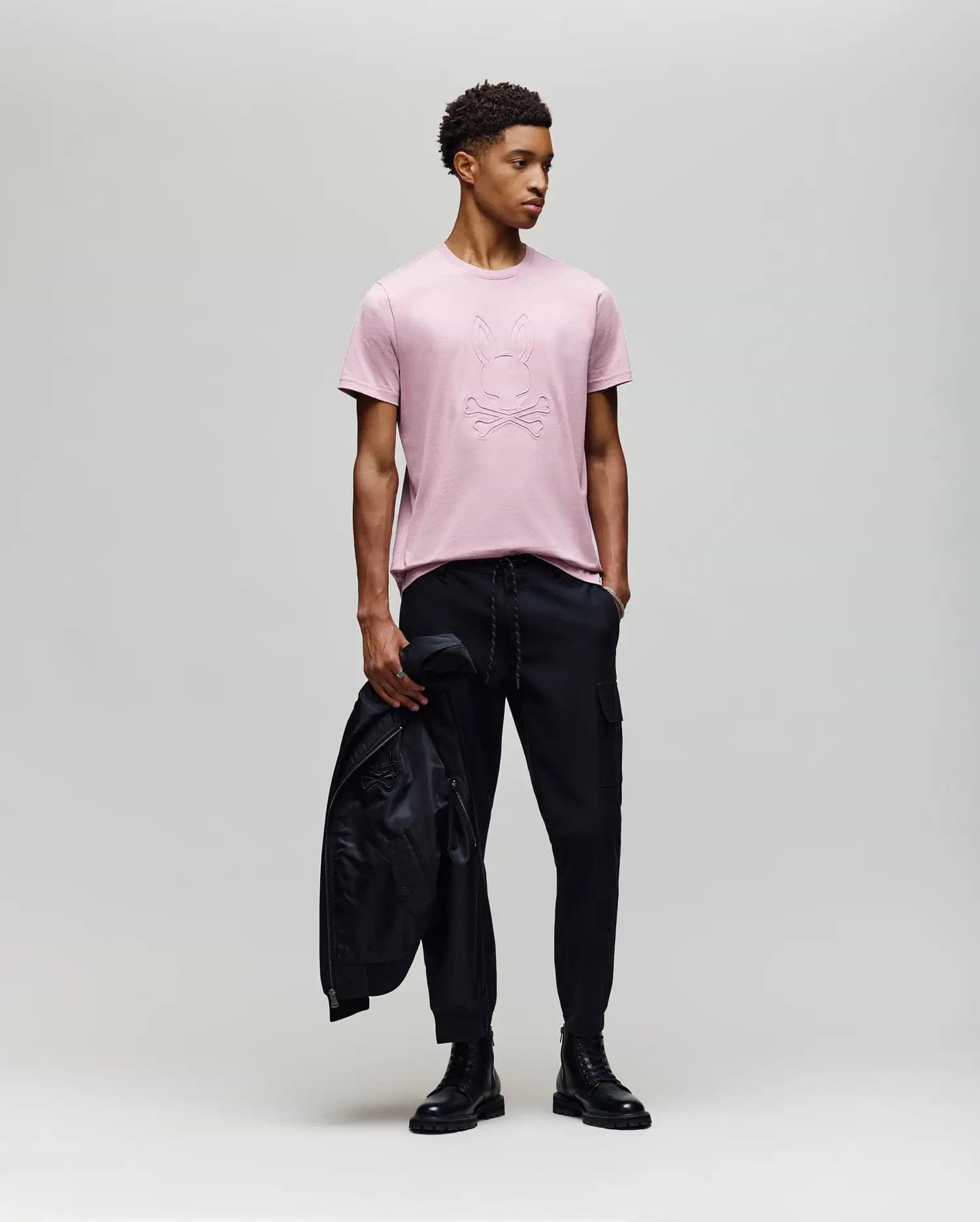 A young man stands against a plain background wearing a light pink Psycho Bunny MENS REMY GRAPHIC TEE - MT0200059 made from soft Pima cotton, paired with black pants and boots, holding a black jacket in his left hand while looking right. (5)