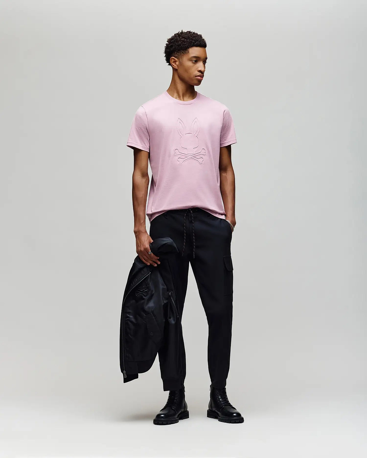 A young man stands against a plain background wearing a light pink Psycho Bunny MENS REMY GRAPHIC TEE - MT0200059 made from soft Pima cotton, paired with black pants and boots, holding a black jacket in his left hand while looking right. (5)
