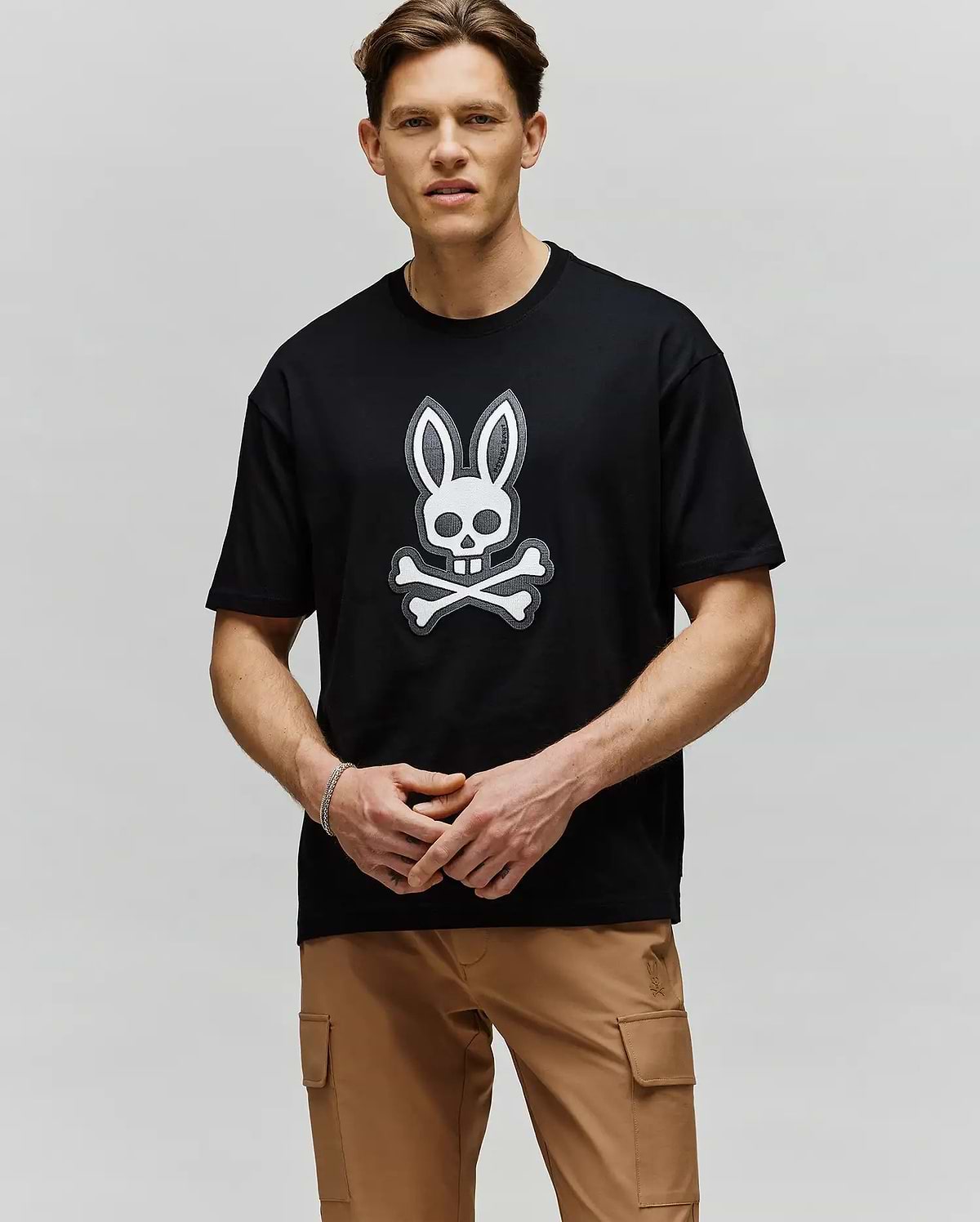A man with short brown hair stands against a light gray background, wearing tan cargo pants and the Psycho Bunny MENS RYDER OVERSIZED FIT TEE (MT0200062) in black, featuring premium Pima cotton and an embroidered Bunny skull graphic.