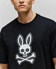 A person wears the Psycho Bunny MENS RYDER OVERSIZED FIT TEE (MT0200062), a black premium shirt featuring a bold white bunny skull and crossbones graphic on the front; only the lower half of their face is shown.