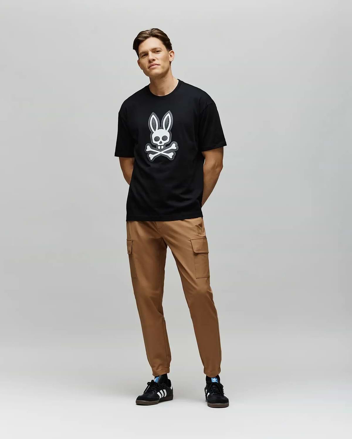 A man with short hair and a relaxed posture wears the Psycho Bunny MENS RYDER OVERSIZED FIT TEE (MT0200062), featuring premium Pima cotton and an embroidered Bunny skull, paired with tan cargo pants and black Adidas sneakers.