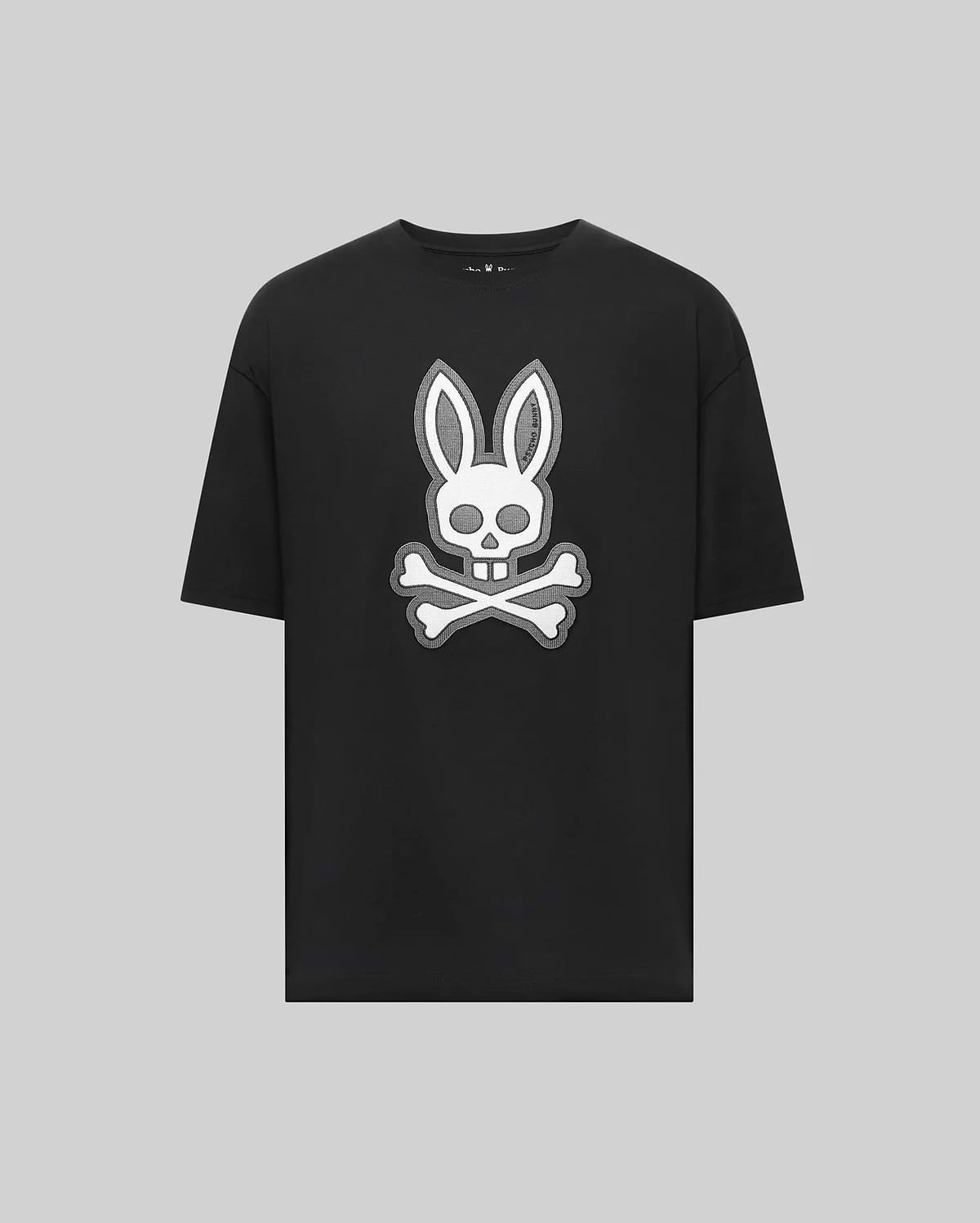 The Psycho Bunny MENS RYDER OVERSIZED FIT TEE (MT0200062) is a black Pima cotton shirt featuring a bold white bunny head with long ears over a skull and crossbones, all set on a plain gray background.