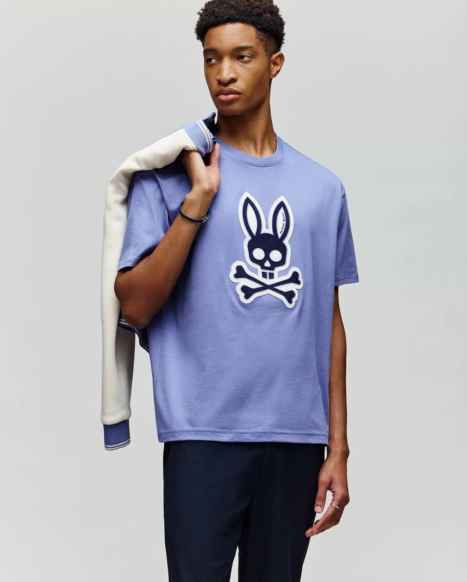 A young man stands in front of a plain light background, holding a jacket and wearing the Psycho Bunny MENS RYDER OVERSIZED FIT TEE (MT0200062), a loose Pima cotton shirt with a bold bunny skull and crossbones graphic, paired with dark pants. (2)
