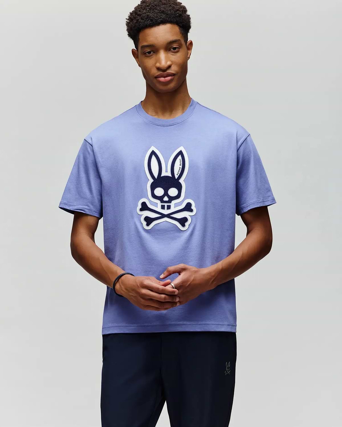 A young man wears the Psycho Bunny MENS RYDER OVERSIZED FIT TEE (MT0200062), a soft Pima cotton shirt with a large cartoon skull and crossbones topped with bunny ears, standing against a plain light background. (4)