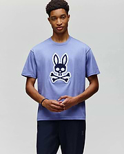 A young man wears the Psycho Bunny MENS RYDER OVERSIZED FIT TEE (MT0200062), a soft Pima cotton shirt with a large cartoon skull and crossbones topped with bunny ears, standing against a plain light background. (4)