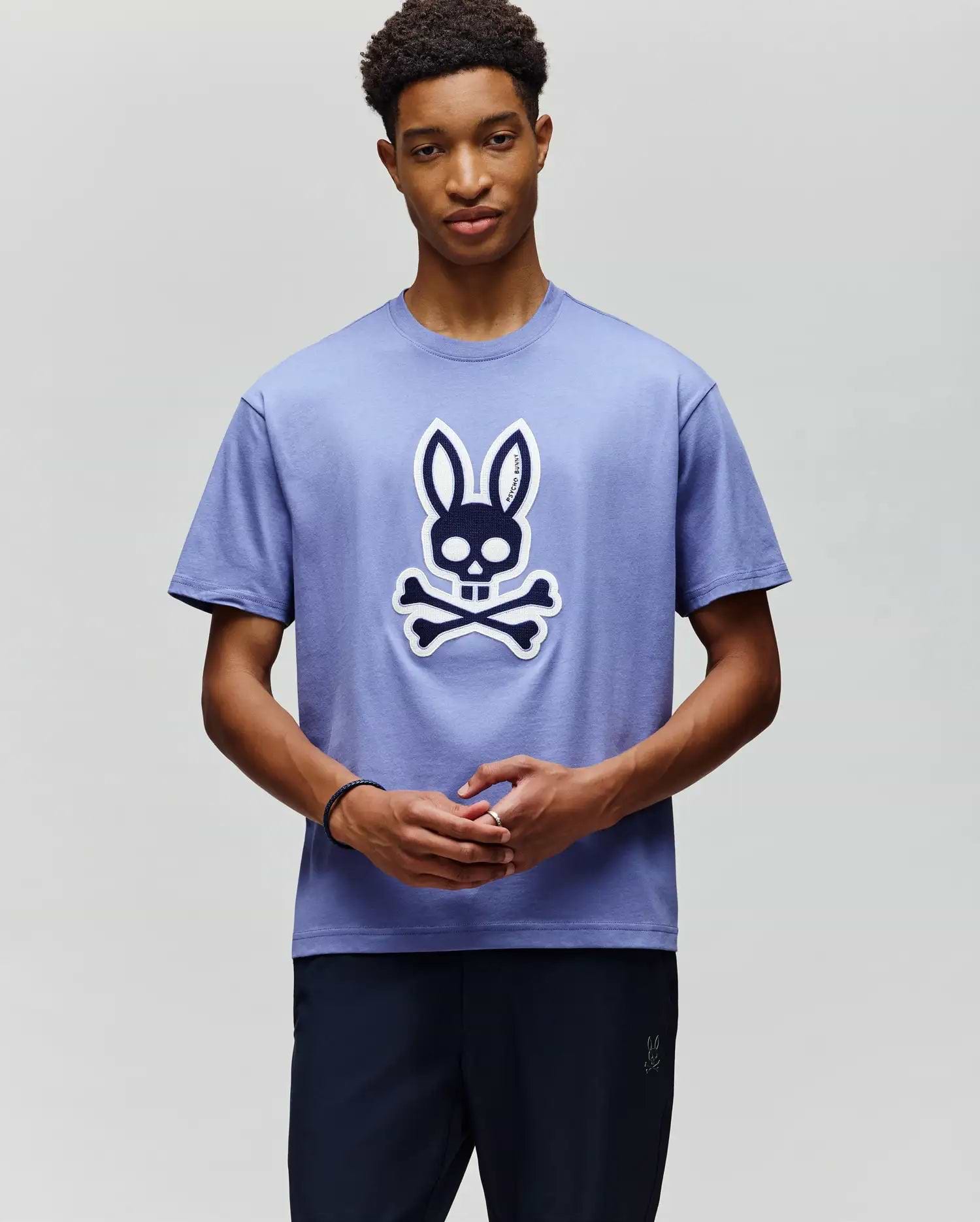 A young man wears the Psycho Bunny MENS RYDER OVERSIZED FIT TEE (MT0200062), a soft Pima cotton shirt with a large cartoon skull and crossbones topped with bunny ears, standing against a plain light background. (4)
