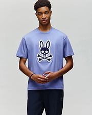 A young man wears the Psycho Bunny MENS RYDER OVERSIZED FIT TEE (MT0200062), a soft Pima cotton shirt with a large cartoon skull and crossbones topped with bunny ears, standing against a plain light background. (4)