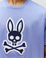 A person wears the Psycho Bunny MENS RYDER OVERSIZED FIT TEE (MT0200062), a light purple premium Pima cotton shirt featuring a bold graphic bunny with skull and crossbones, outlined ears, and facial details in white and black. (3)