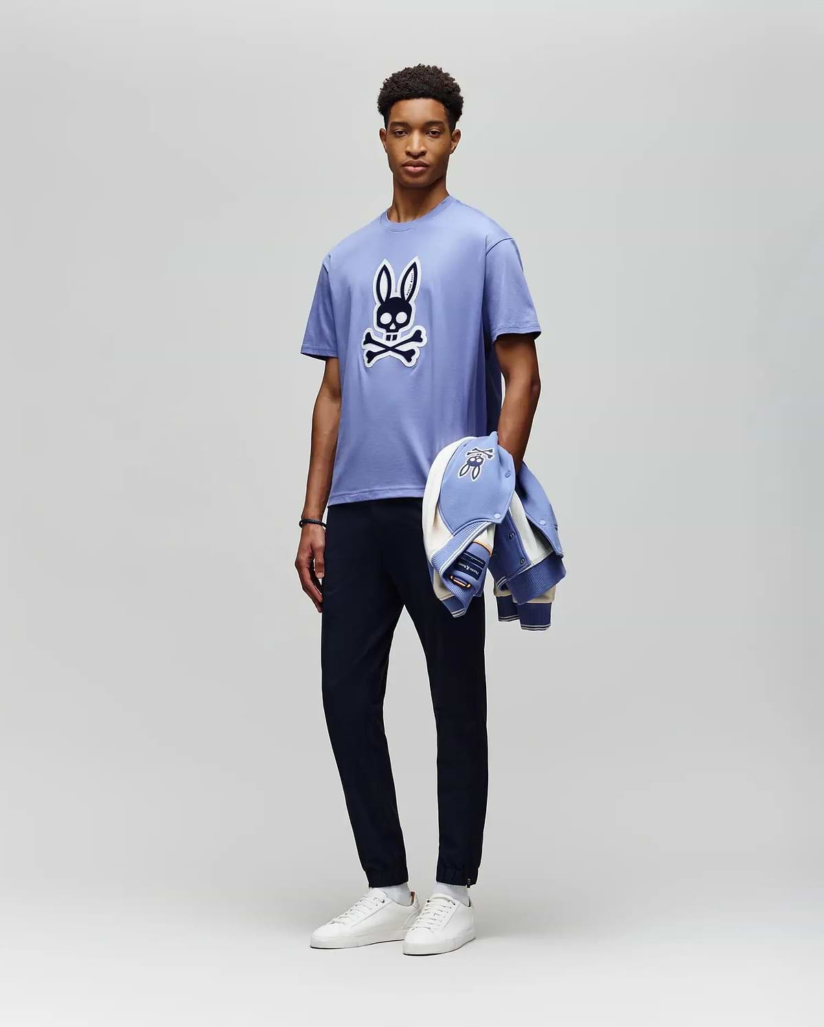 A man stands against a plain background wearing the Psycho Bunny MENS RYDER OVERSIZED FIT TEE (MT0200062), a light purple premium Pima cotton shirt with an embroidered Bunny skull and crossbones, dark pants, white sneakers, and holds a matching jacket. (5)