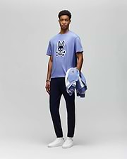A man stands against a plain background wearing the Psycho Bunny MENS RYDER OVERSIZED FIT TEE (MT0200062), a light purple premium Pima cotton shirt with an embroidered Bunny skull and crossbones, dark pants, white sneakers, and holds a matching jacket. (5)