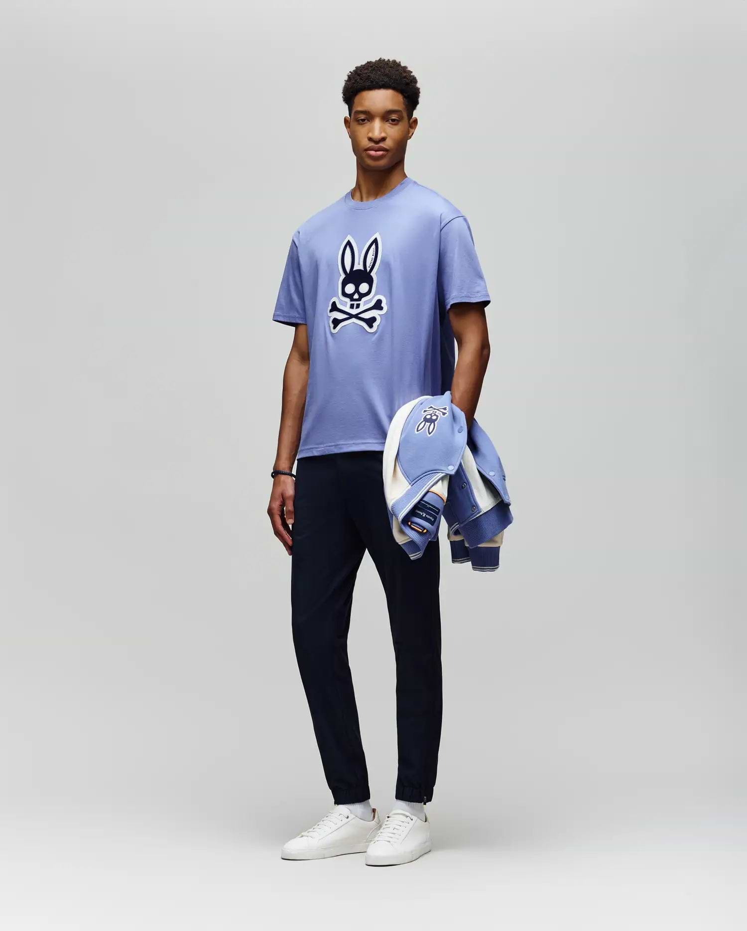 A man stands against a plain background wearing the Psycho Bunny MENS RYDER OVERSIZED FIT TEE (MT0200062), a light purple premium Pima cotton shirt with an embroidered Bunny skull and crossbones, dark pants, white sneakers, and holds a matching jacket. (5)