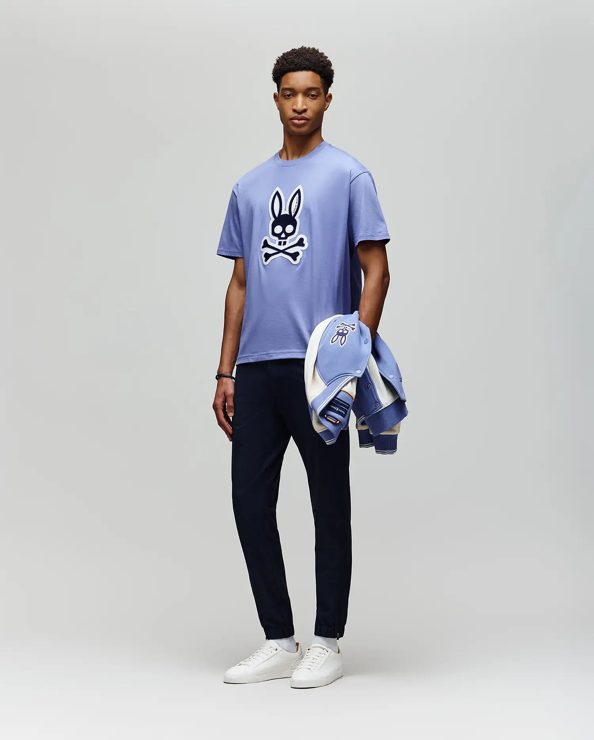 A man stands against a plain background wearing the Psycho Bunny MENS RYDER OVERSIZED FIT TEE (MT0200062), a light purple premium Pima cotton shirt with an embroidered Bunny skull and crossbones, dark pants, white sneakers, and holds a matching jacket. (5)