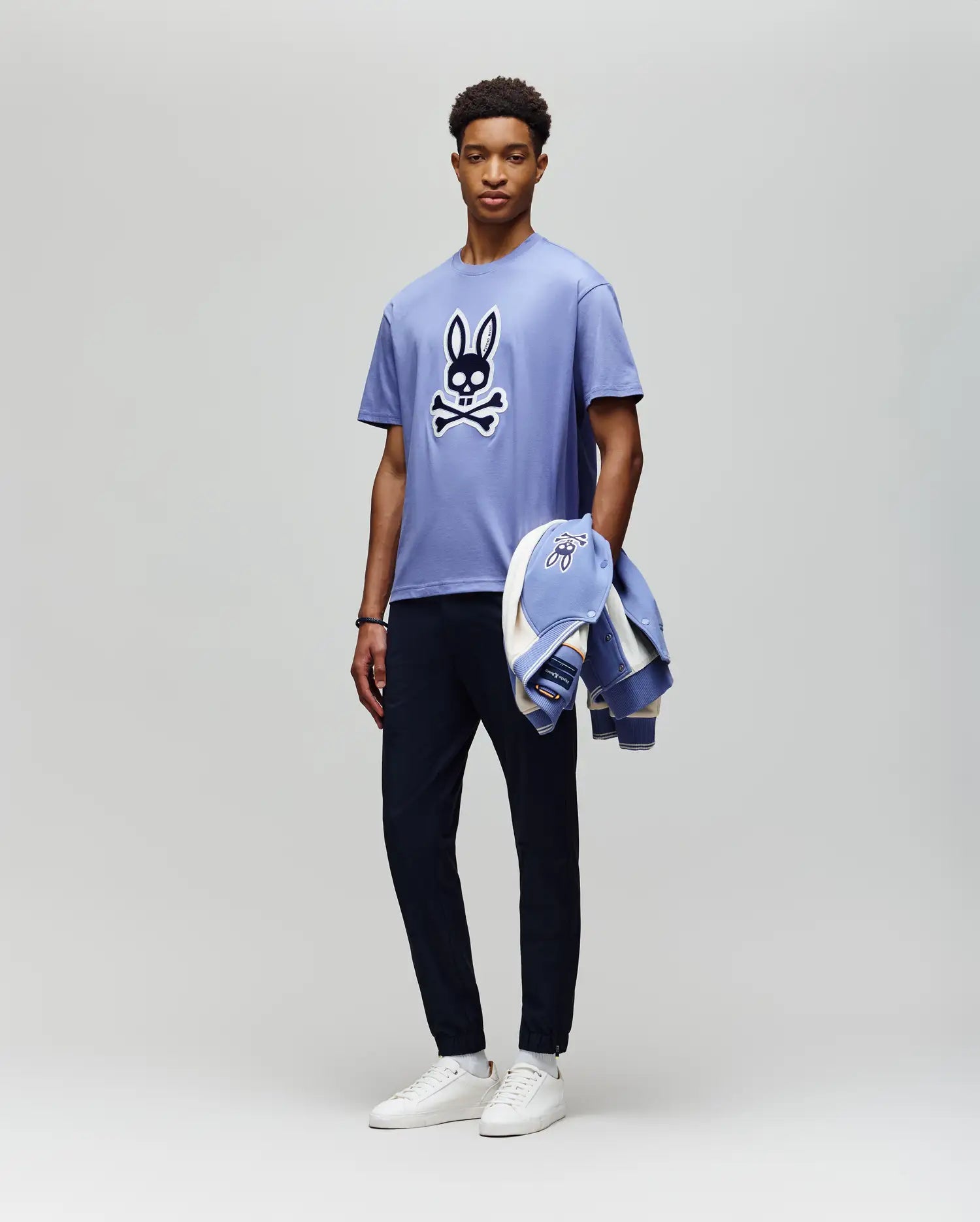 A man stands against a plain background wearing the Psycho Bunny MENS RYDER OVERSIZED FIT TEE (MT0200062), a light purple premium Pima cotton shirt with an embroidered Bunny skull and crossbones, dark pants, white sneakers, and holds a matching jacket. (5)
