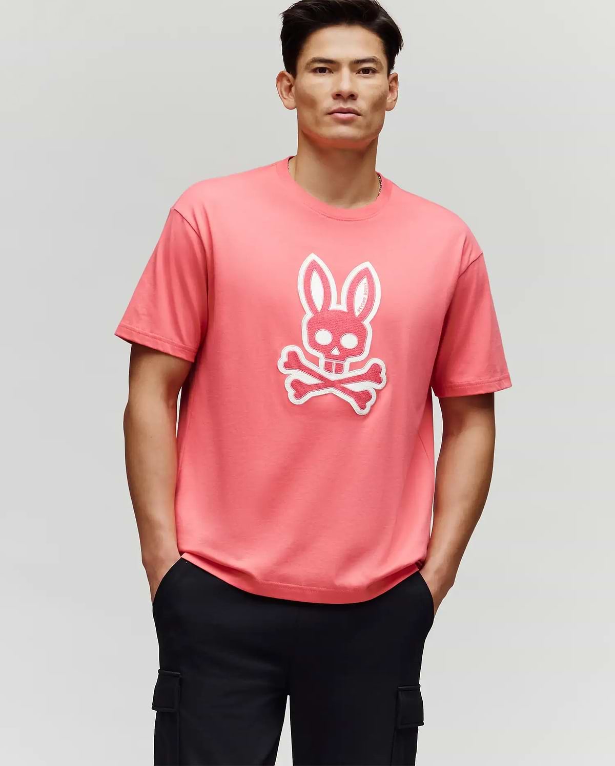 A man stands with his hands in his black pants pockets, wearing the Psycho Bunny MENS RYDER OVERSIZED FIT TEE (MT0200062), a pink premium tee featuring a white bunny skull and crossbones graphic, against a plain light gray background.