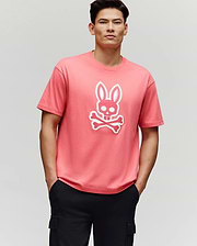 A man stands with his hands in his black pants pockets, wearing the Psycho Bunny MENS RYDER OVERSIZED FIT TEE (MT0200062), a pink premium tee featuring a white bunny skull and crossbones graphic, against a plain light gray background.