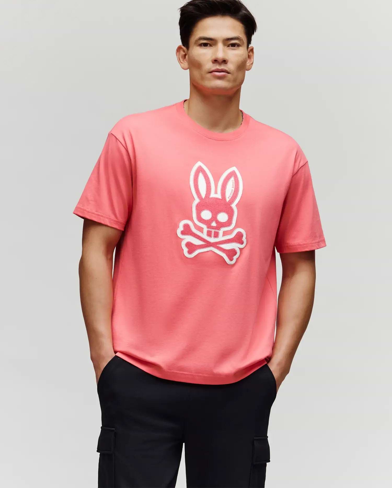 A man stands with his hands in his black pants pockets, wearing the Psycho Bunny MENS RYDER OVERSIZED FIT TEE (MT0200062), a pink premium tee featuring a white bunny skull and crossbones graphic, against a plain light gray background.