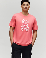 A man stands with his hands in his black pants pockets, wearing the Psycho Bunny MENS RYDER OVERSIZED FIT TEE (MT0200062), a pink premium tee featuring a white bunny skull and crossbones graphic, against a plain light gray background.