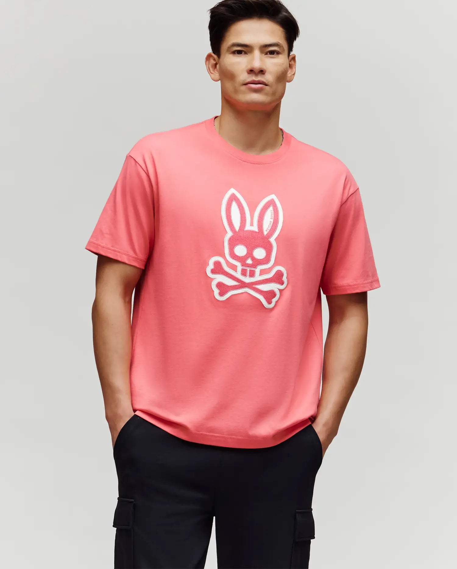 A man stands with his hands in his black pants pockets, wearing the Psycho Bunny MENS RYDER OVERSIZED FIT TEE (MT0200062), a pink premium tee featuring a white bunny skull and crossbones graphic, against a plain light gray background.