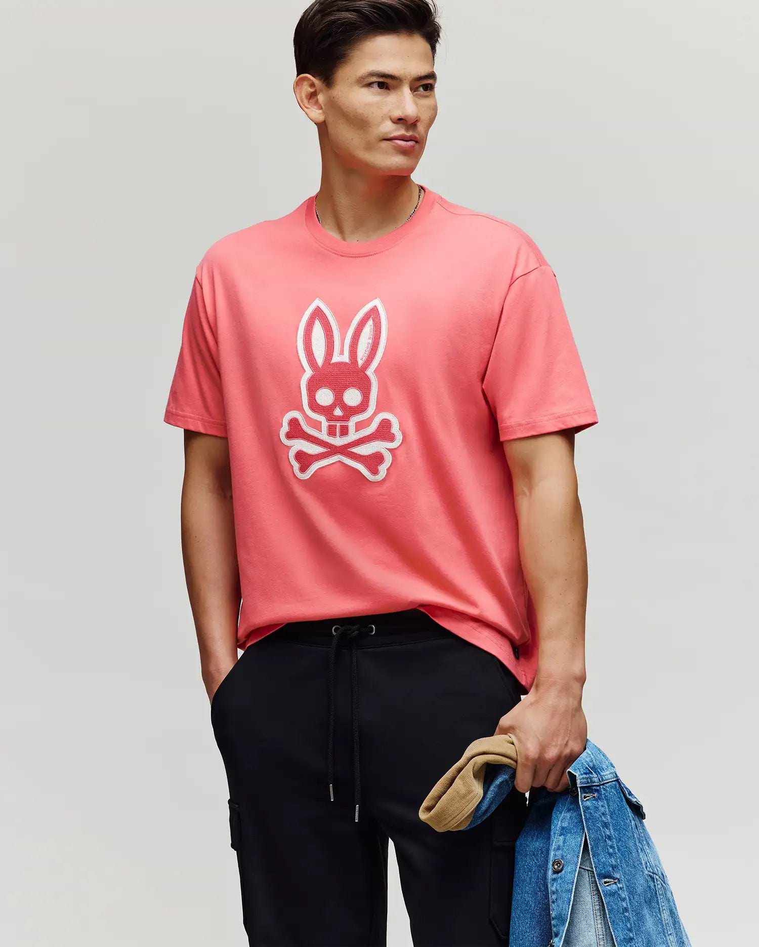A man wears the Psycho Bunny MENS RYDER OVERSIZED FIT TEE (MT0200062) in pink, featuring an embroidered Bunny skull and crossbones. He pairs it with black pants, holds a denim jacket and tan cap, and stands with a hand in his pocket.