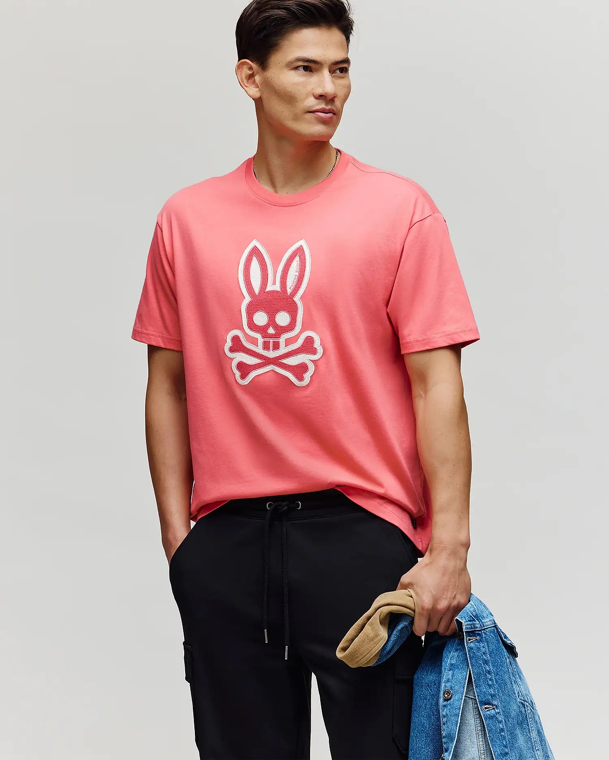 A man wears the Psycho Bunny MENS RYDER OVERSIZED FIT TEE (MT0200062) in pink, featuring an embroidered Bunny skull and crossbones. He pairs it with black pants, holds a denim jacket and tan cap, and stands with a hand in his pocket.