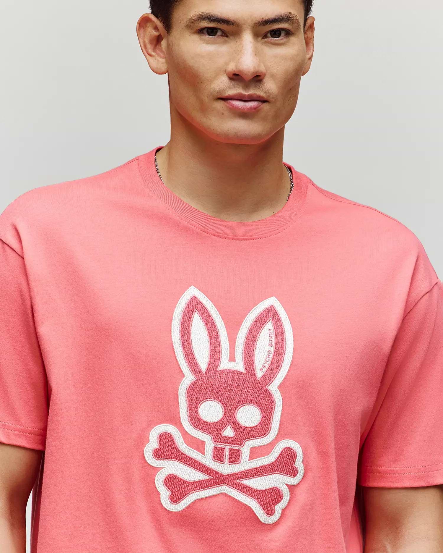 A person wears the Psycho Bunny MENS RYDER OVERSIZED FIT TEE (MT0200062) in pink, featuring a large embroidered Bunny skull and crossbones graphic on the front, standing against a plain light background.