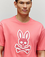 A person wears the Psycho Bunny MENS RYDER OVERSIZED FIT TEE (MT0200062) in pink, featuring a large embroidered Bunny skull and crossbones graphic on the front, standing against a plain light background.