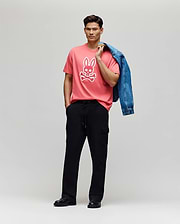 A man stands against a plain backdrop wearing the Psycho Bunny MENS RYDER OVERSIZED FIT TEE - MT0200062, crafted from soft Pima cotton with an embroidered Bunny, paired with black cargo pants, black shoes, and holding a blue denim jacket.