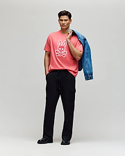 A man stands against a plain backdrop wearing the Psycho Bunny MENS RYDER OVERSIZED FIT TEE - MT0200062, crafted from soft Pima cotton with an embroidered Bunny, paired with black cargo pants, black shoes, and holding a blue denim jacket.