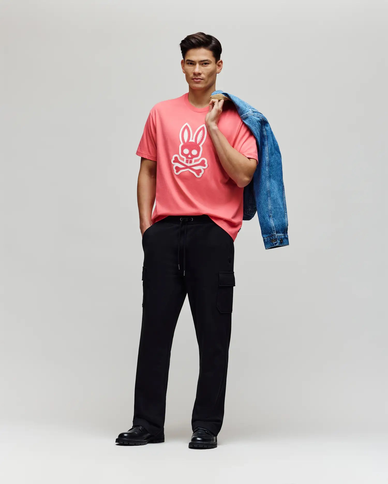 A man stands against a plain backdrop wearing the Psycho Bunny MENS RYDER OVERSIZED FIT TEE - MT0200062, crafted from soft Pima cotton with an embroidered Bunny, paired with black cargo pants, black shoes, and holding a blue denim jacket.