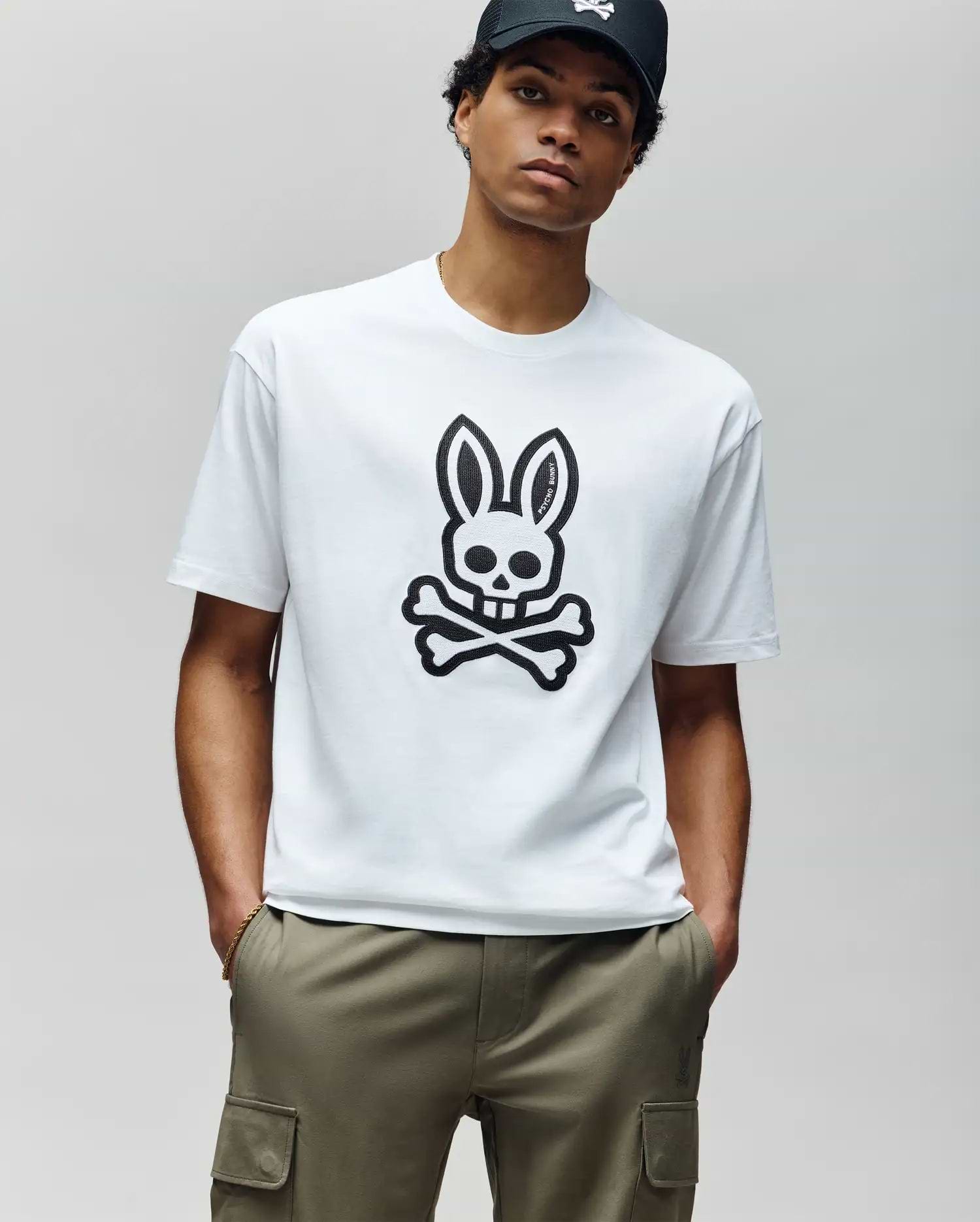 A man in a black cap, olive green cargo pants, and a Psycho Bunny MENS RYDER OVERSIZED FIT TEE—featuring an embroidered Bunny skull and crossbones—stands with hands in pockets against a plain light gray background.