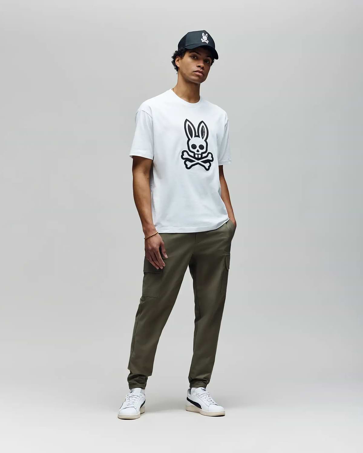 A man stands against a plain background wearing the Psycho Bunny MENS RYDER OVERSIZED FIT TEE (MT0200062) with a large black skull and embroidered Bunny graphic, olive green pants, white sneakers, and a black cap. His hands are in his pockets.