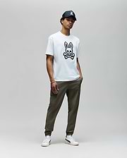 A man stands against a plain background wearing the Psycho Bunny MENS RYDER OVERSIZED FIT TEE (MT0200062) with a large black skull and embroidered Bunny graphic, olive green pants, white sneakers, and a black cap. His hands are in his pockets.
