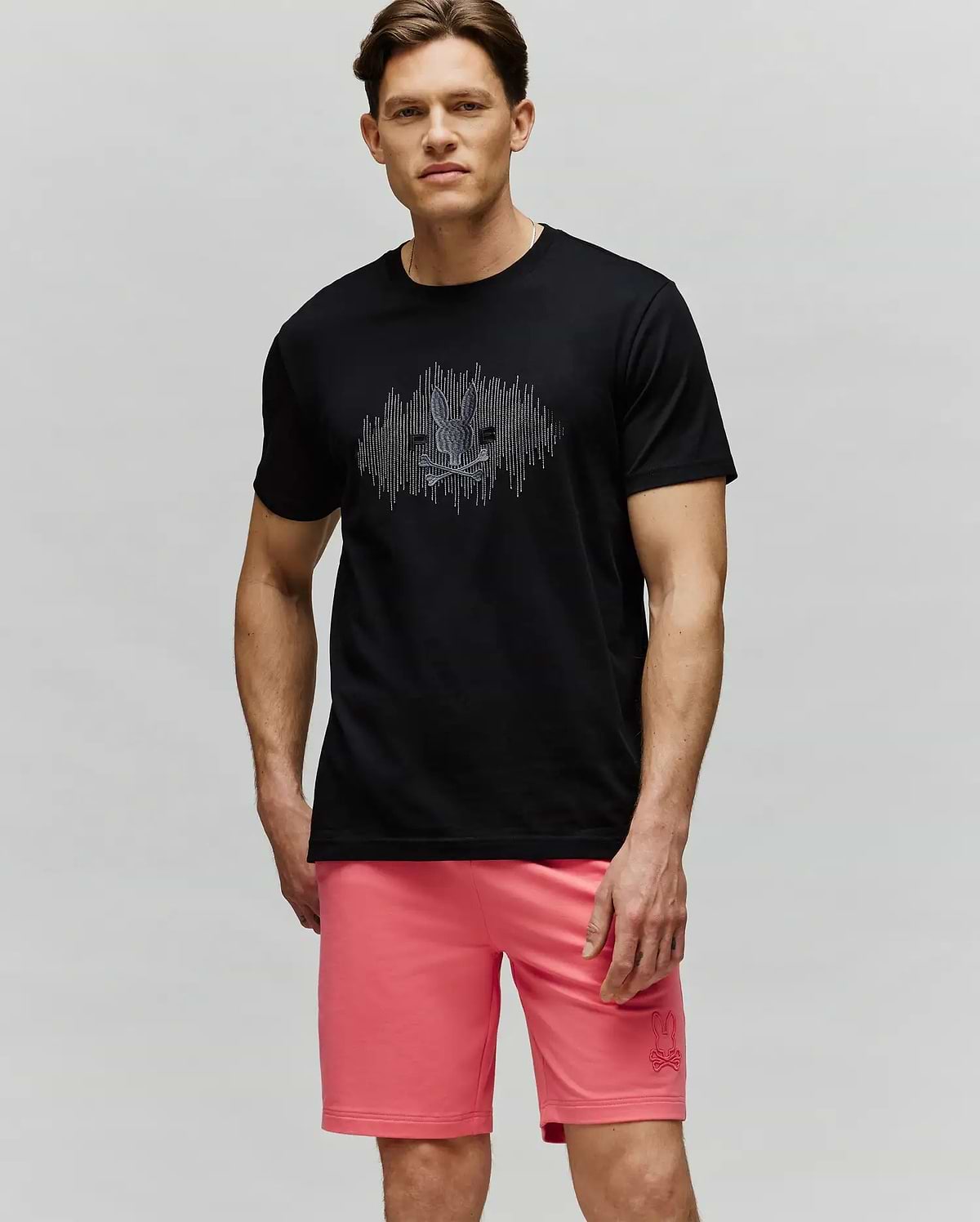 A man with short brown hair and a neutral expression wears the Psycho Bunny MENS OLLIE GRAPHIC TEE (MT0200066) in black, featuring a bunny graphic, paired with pink shorts, standing against a light background. (4)