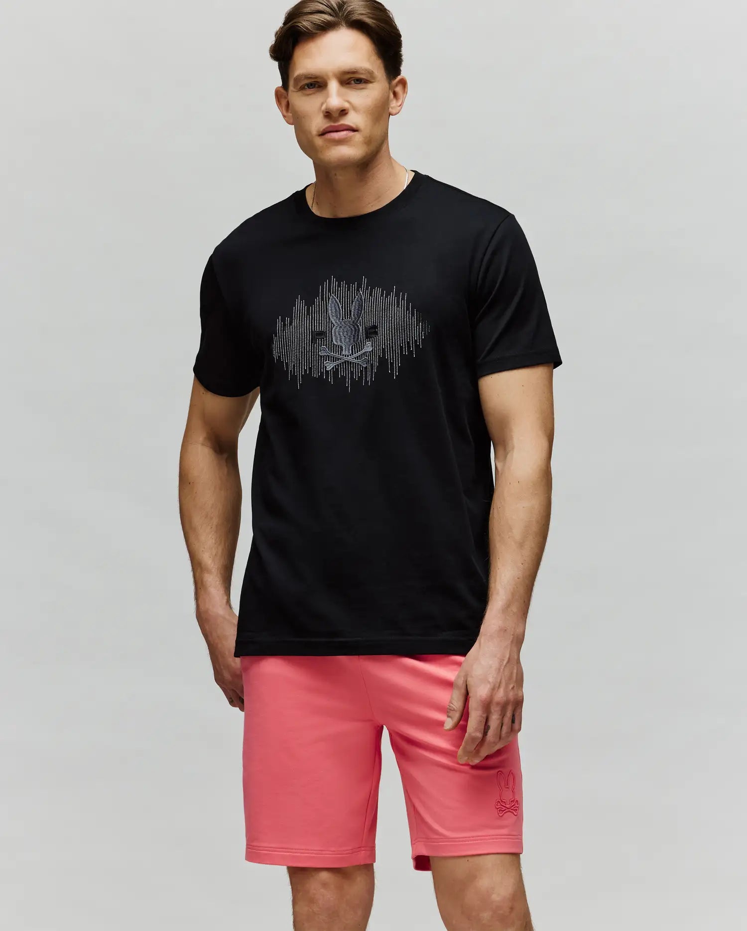 A man with short brown hair and a neutral expression wears the Psycho Bunny MENS OLLIE GRAPHIC TEE (MT0200066) in black, featuring a bunny graphic, paired with pink shorts, standing against a light background. (4)
