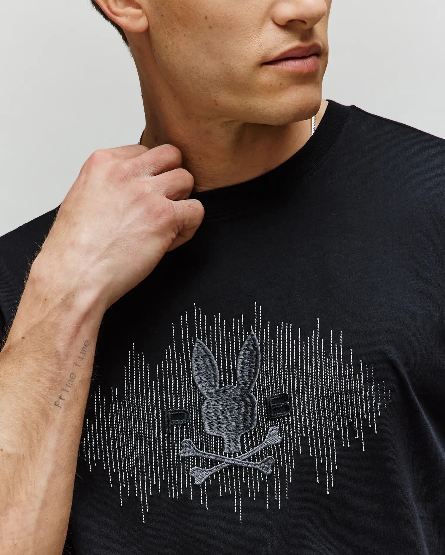 A man wears the Psycho Bunny MENS OLLIE GRAPHIC TEE - MT0200066 in black, showcasing its embroidered skull, crossbones, and bunny graphic. His raised hand highlights a faint forearm tattoo; the photo is cropped at his shoulders. (3)