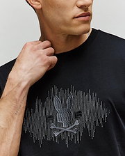 A man wears the Psycho Bunny MENS OLLIE GRAPHIC TEE - MT0200066 in black, showcasing its embroidered skull, crossbones, and bunny graphic. His raised hand highlights a faint forearm tattoo; the photo is cropped at his shoulders. (3)