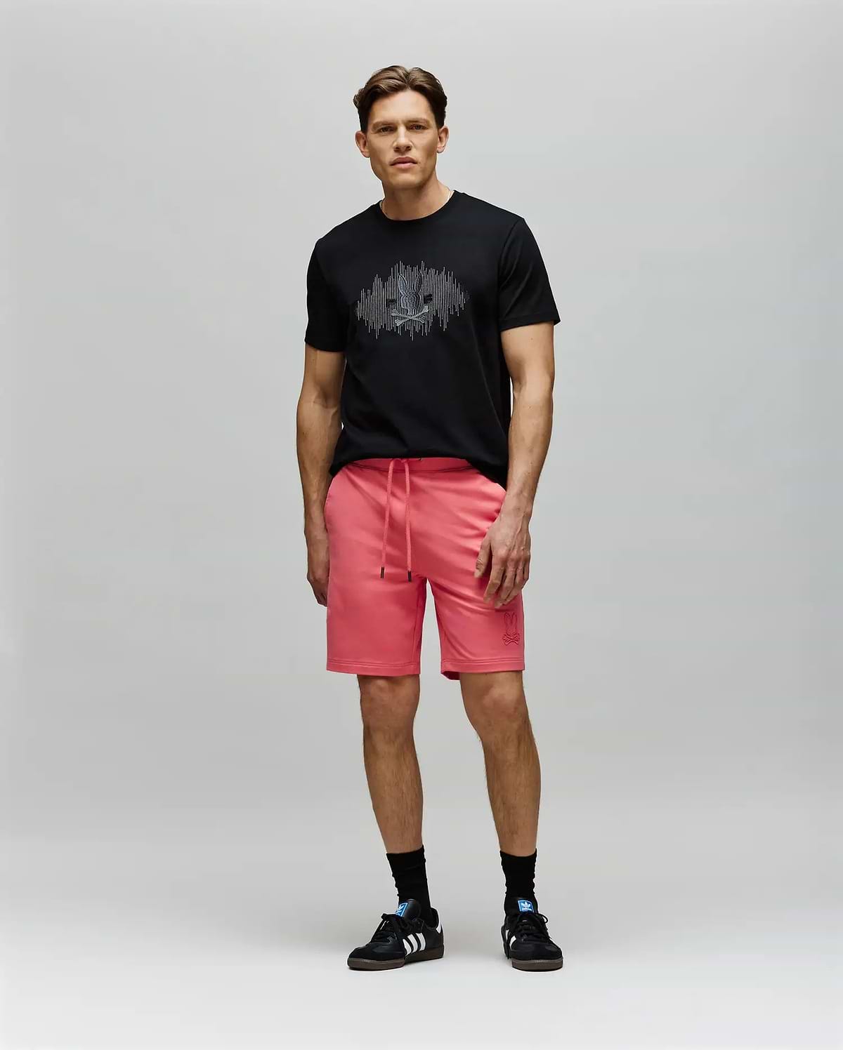 A man stands against a light gray background wearing a black Psycho Bunny MENS OLLIE GRAPHIC TEE (MT0200066) with a Bunny graphic, bright pink drawstring shorts, black socks, and black Adidas sneakers, looking forward neutrally. (5)