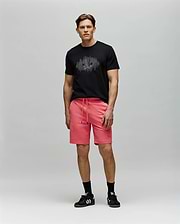 A man stands against a light gray background wearing a black Psycho Bunny MENS OLLIE GRAPHIC TEE (MT0200066) with a Bunny graphic, bright pink drawstring shorts, black socks, and black Adidas sneakers, looking forward neutrally. (5)