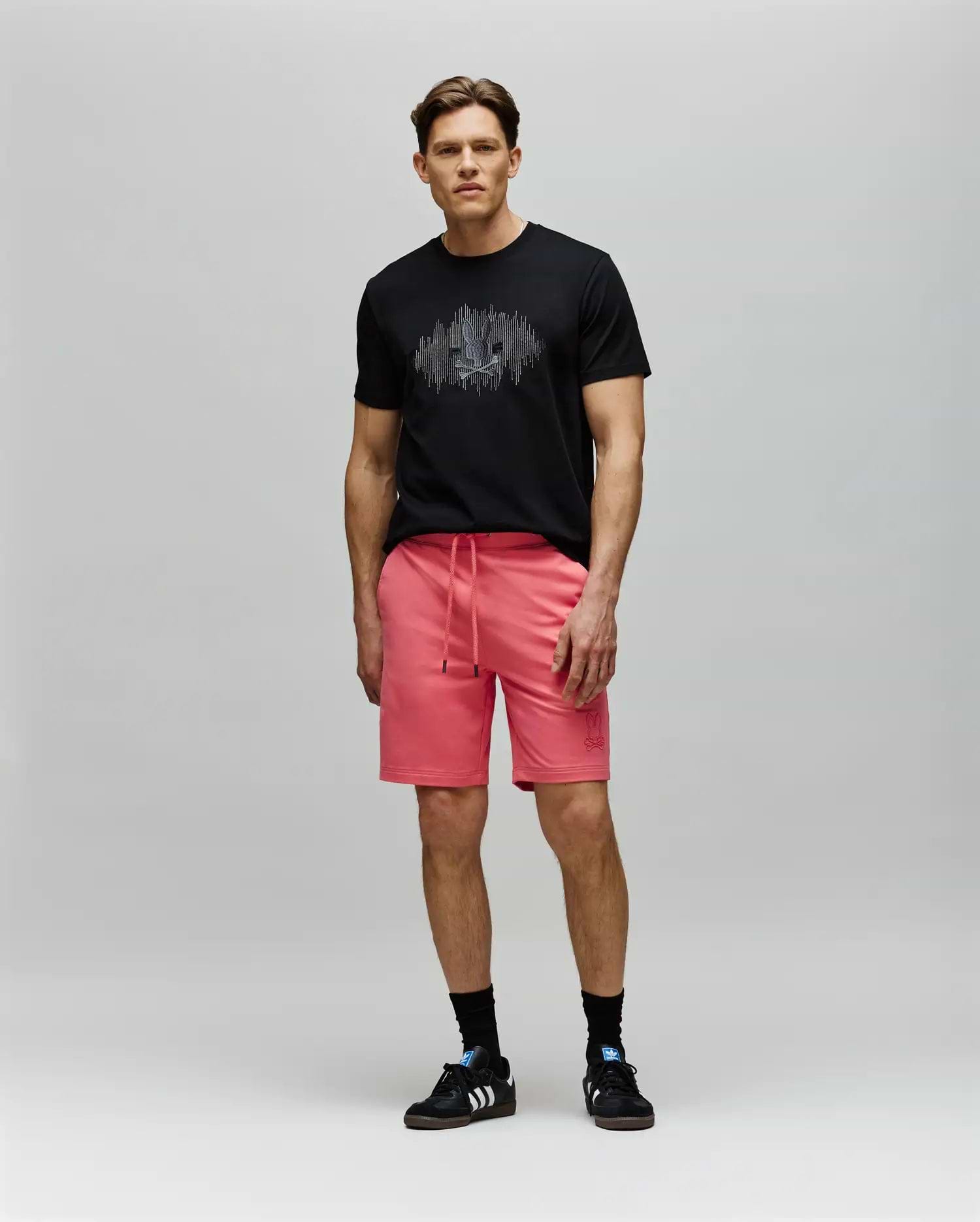 A man stands against a light gray background wearing a black Psycho Bunny MENS OLLIE GRAPHIC TEE (MT0200066) with a Bunny graphic, bright pink drawstring shorts, black socks, and black Adidas sneakers, looking forward neutrally. (5)