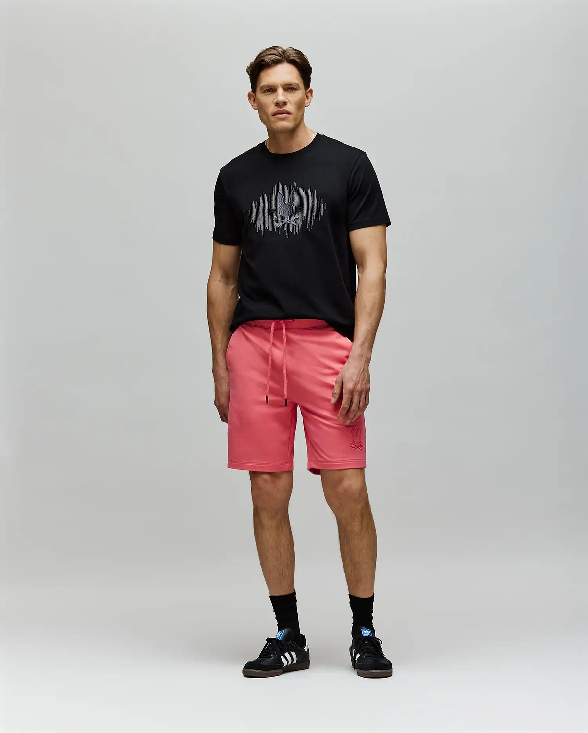 A man stands against a light gray background wearing a black Psycho Bunny MENS OLLIE GRAPHIC TEE (MT0200066) with a Bunny graphic, bright pink drawstring shorts, black socks, and black Adidas sneakers, looking forward neutrally. (5)