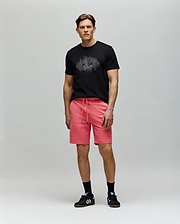 A man stands against a light gray background wearing a black Psycho Bunny MENS OLLIE GRAPHIC TEE (MT0200066) with a Bunny graphic, bright pink drawstring shorts, black socks, and black Adidas sneakers, looking forward neutrally. (5)