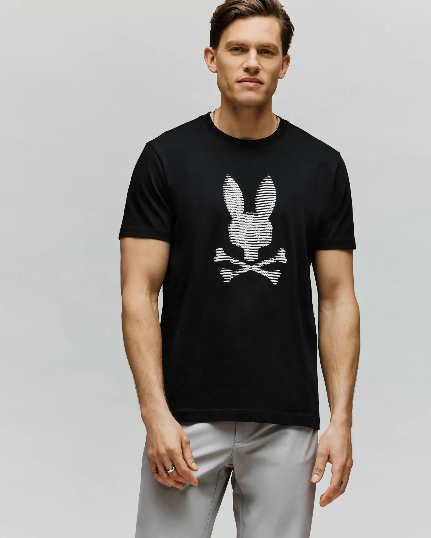 A man wears the Psycho Bunny MENS GRANT BUNNY GRAPHIC TEE - MT0200081, featuring an HD-printed white bunny head above crossed bones. He stands against a plain light background, short-haired and looking at the camera with a neutral expression.