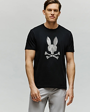 A man wears the Psycho Bunny MENS GRANT BUNNY GRAPHIC TEE - MT0200081, featuring an HD-printed white bunny head above crossed bones. He stands against a plain light background, short-haired and looking at the camera with a neutral expression.