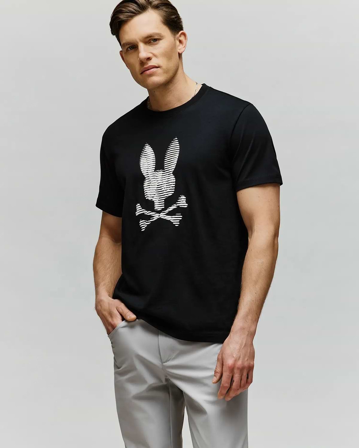 A man with short light brown hair wears Psycho Bunny’s MENS GRANT BUNNY GRAPHIC TEE (MT0200081)—a black tee with an HD-printed white bunny head and crossbones—paired with light gray pants, posing with one hand in his pocket.