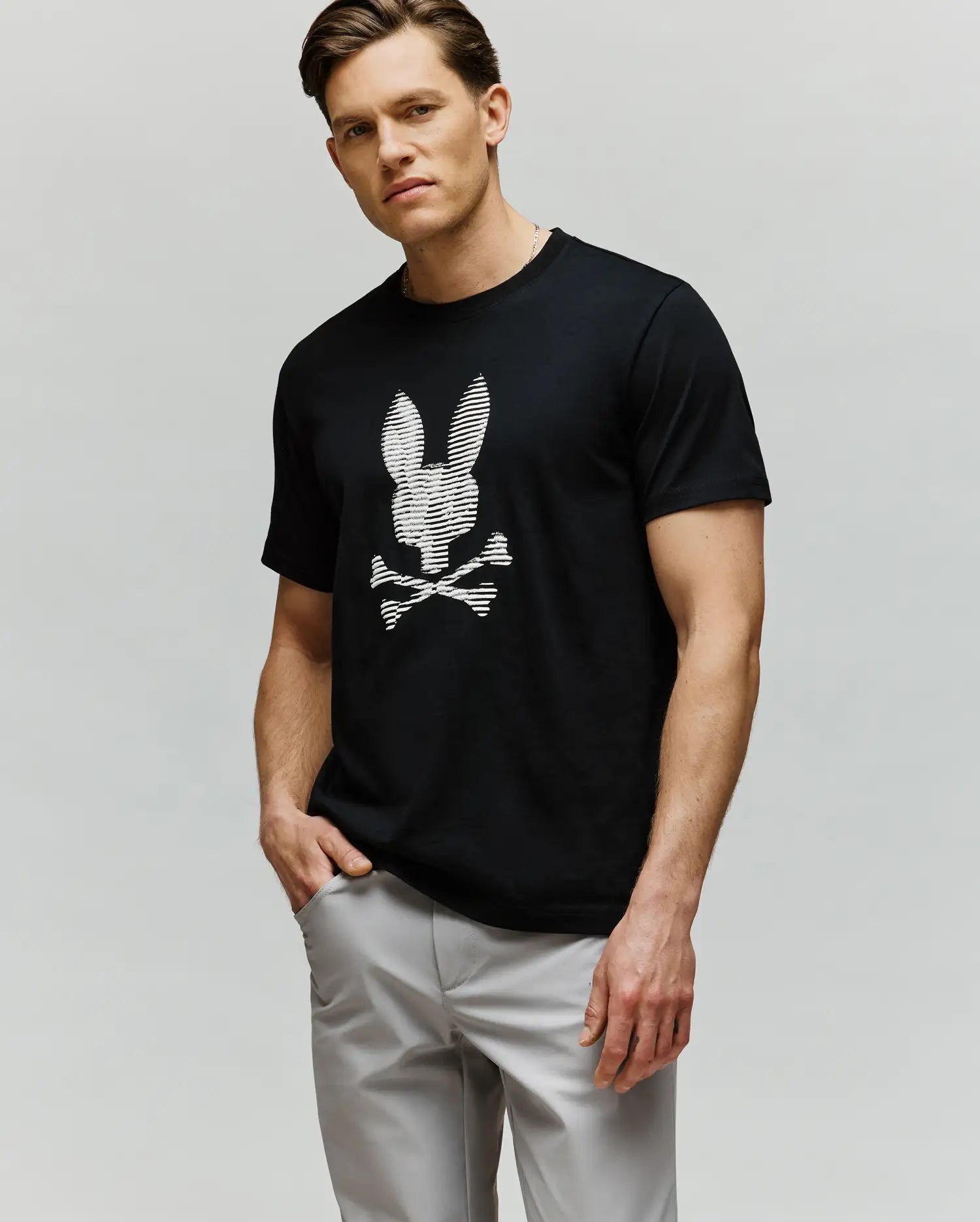 A man with short light brown hair wears Psycho Bunny’s MENS GRANT BUNNY GRAPHIC TEE (MT0200081)—a black tee with an HD-printed white bunny head and crossbones—paired with light gray pants, posing with one hand in his pocket.